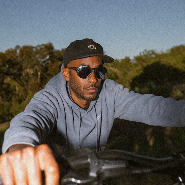 Man wearing sunglasses and a Crankbrothers cap pushing a bicycle with trees in the background